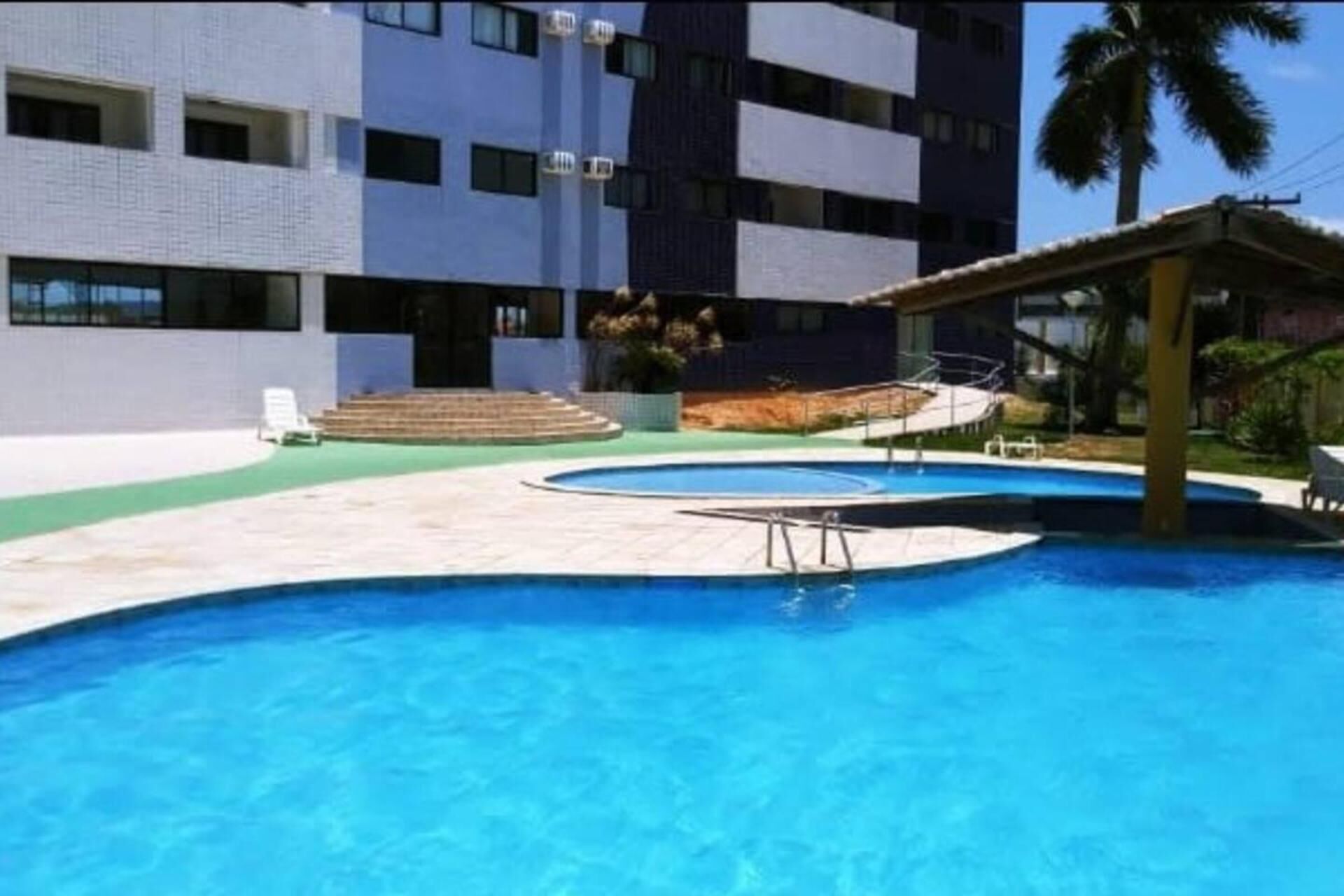Piscina com superfície de água azul cristalina, rodeada por um espaço com pavimento cerâmico. Há degraus que levam à piscina e uma área de estar coberta com telhado.