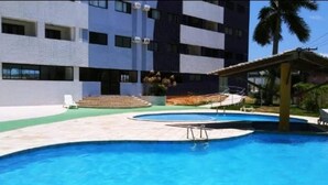 Piscina com superfície de água azul cristalina, rodeada por um espaço com pavimento cerâmico. Há degraus que levam à piscina e uma área de estar coberta com telhado.