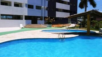 Piscina com superfície de água azul cristalina, rodeada por um espaço com pavimento cerâmico. Há degraus que levam à piscina e uma área de estar coberta com telhado.