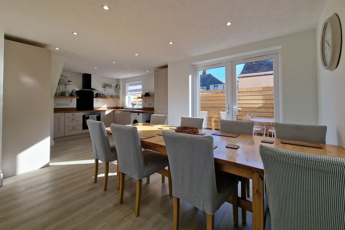 Una cocina moderna con gabinetes blancos, encimera de madera y horno empotrado. Cuenta con una zona de comedor con mesa de madera y sillas a rayas.