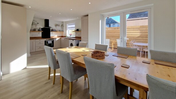 A modern kitchen with white cabinets, a wooden countertop, and a built-in oven. There is a dining area with a wooden table and striped chairs.