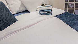 Ein ordentlich bezogenes Bett mit weißen Textilien, einem grauen, gepolsterten Kopfende und einem gefalteten blauen Handtuch.