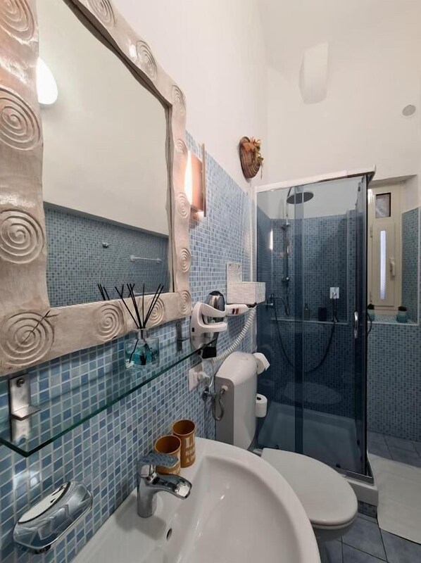 A bathroom with a glass shower enclosure, a white sink, and a mirror with a decorative frame. - Centurions Room (Roma)