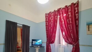 A room with a bed, a TV on a stand, a window with red curtains, and a door leading to another room.