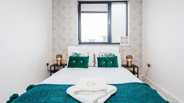 A neatly made bed with a teal blanket and a white towel with a monogram, flanked by two bedside tables with lamps, in a room with a large window and patterned wallpaper. - 2-bed Apartment Near Basildon Train Station (Basildon)