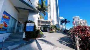 A hotel entrance with a reception desk, palm trees, and modern buildings in the background.