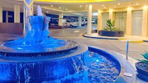 A hotel entrance with a circular fountain, a covered parking area, and potted plants.