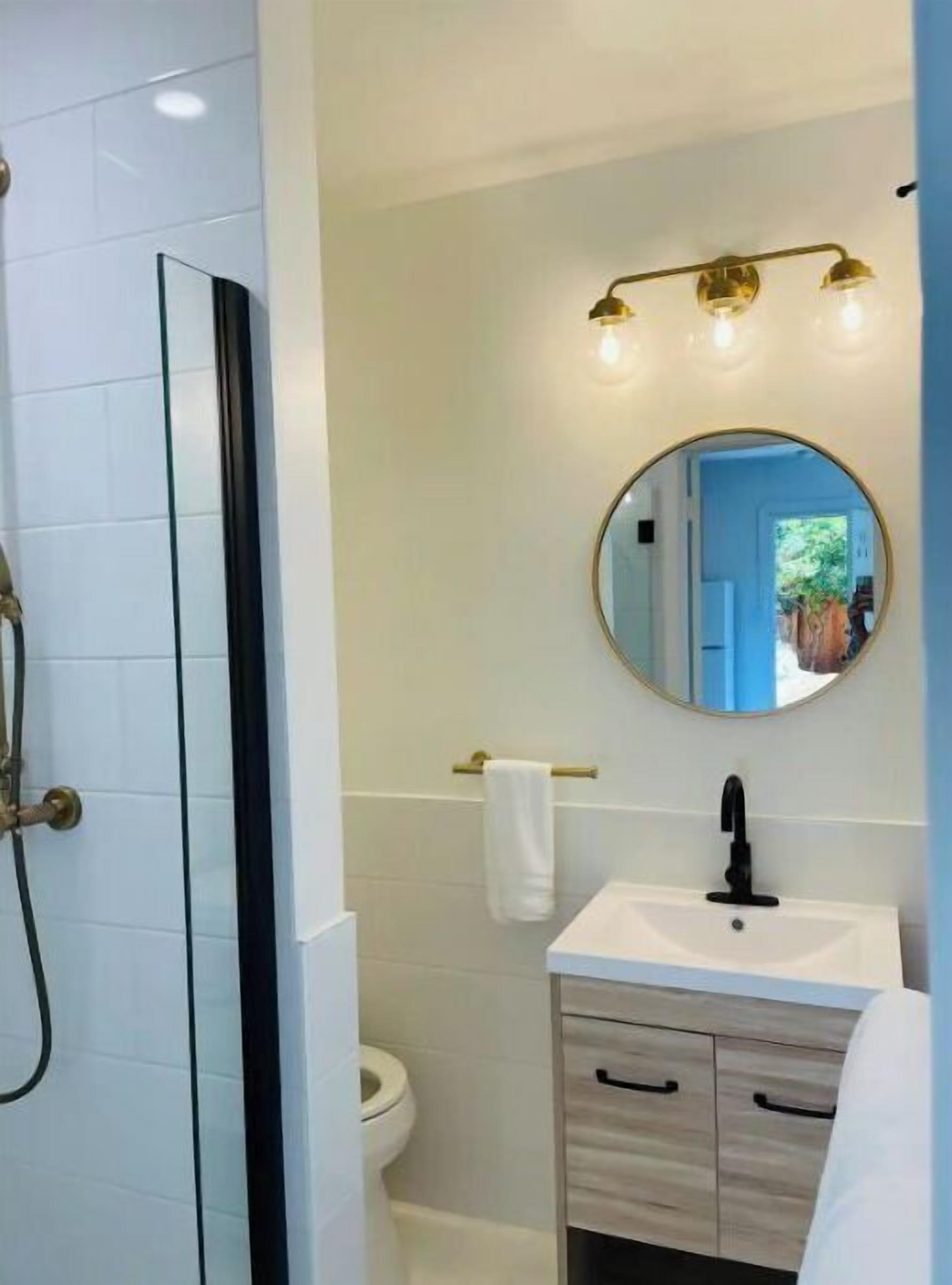 A bathroom with a glass shower enclosure, a white sink with a black faucet, a round mirror with three lights, and a towel rack.