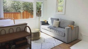 A cozy living room with a grey sofa, a wooden coffee table, and a small round ottoman. There is a window with a view of a wooden fence and greenery outside. - Luxe modern Lagunitas studio at Samuel P Taylor (Lagunitas-Forest Knolls)