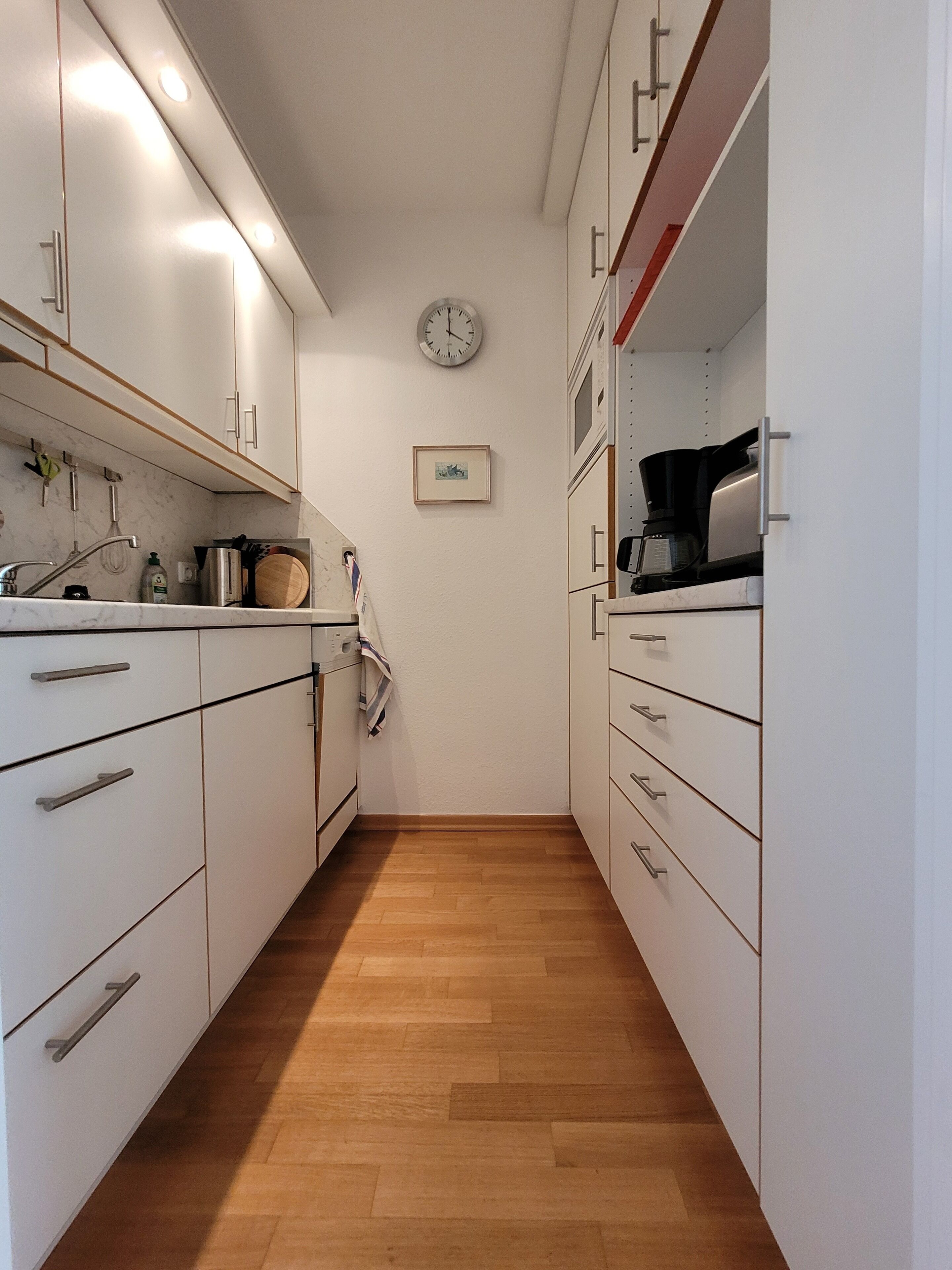 A narrow kitchen with white cabinets, a wooden floor, and a clock on the wall.