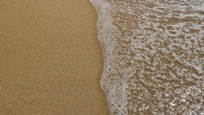 Una playa con olas rompiendo suavemente en la orilla, un cielo despejado y algunas personas a lo lejos.