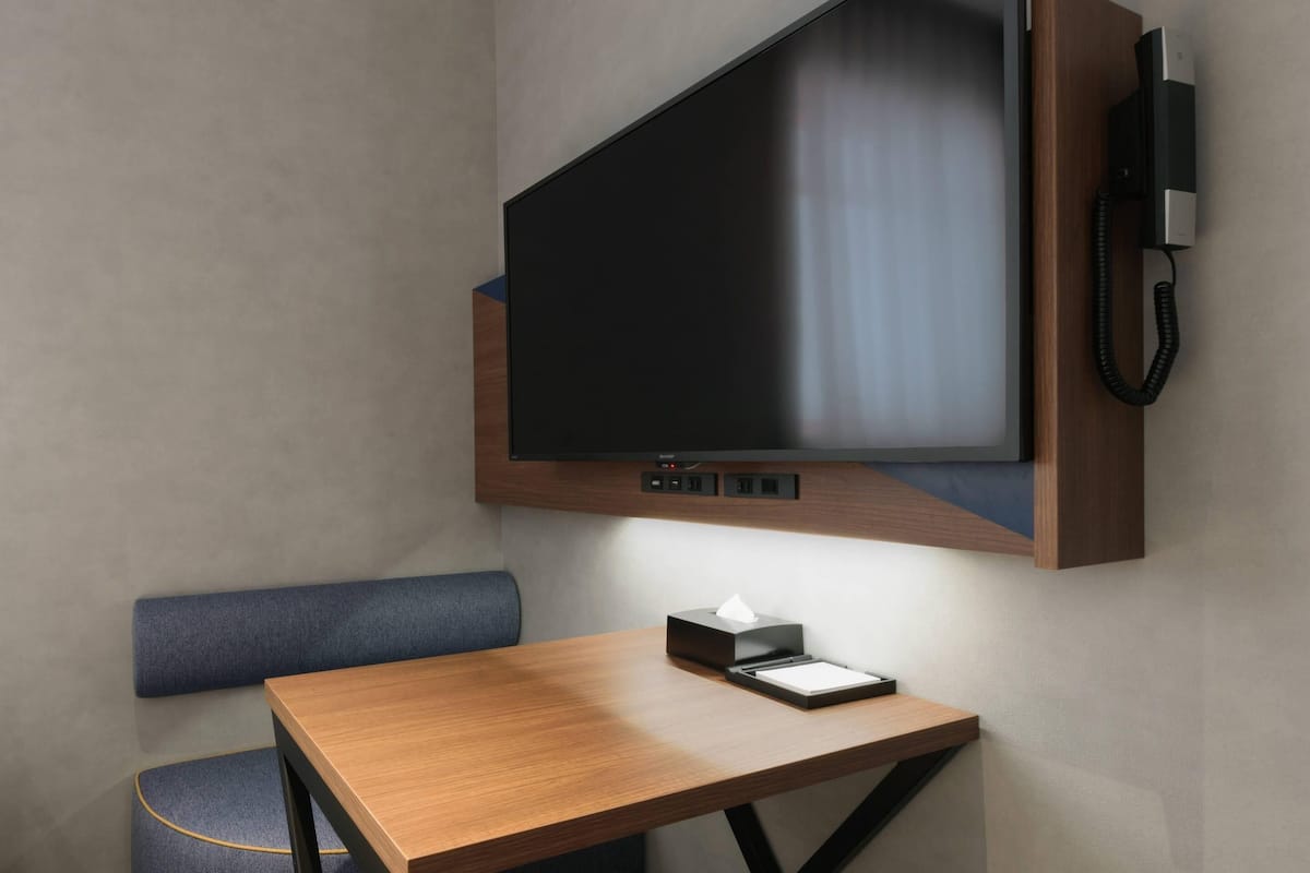 A modern hotel room with a flat-screen TV mounted on the wall, a wooden table with a tissue box, and a blue upholstered bench.