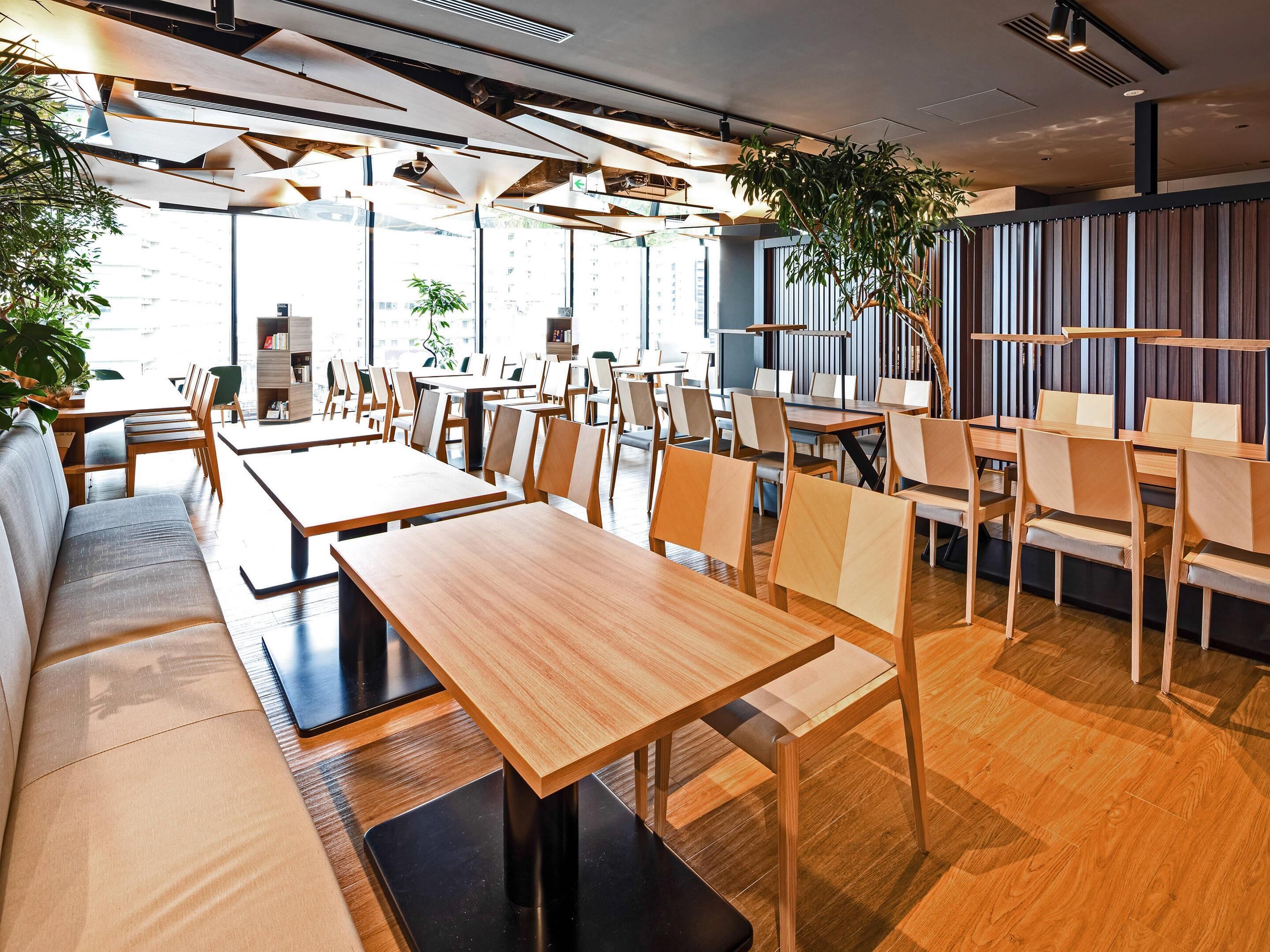 A modern restaurant with wooden tables and chairs, large windows, and indoor plants.