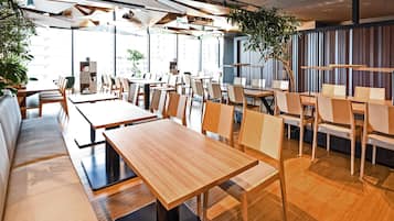 Un restaurante moderno con mesas y sillas de madera, ventanas amplias y plantas de interior.
