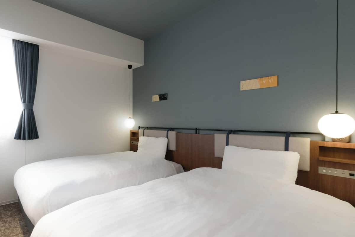 A hotel room with two beds, a nightstand, a lamp, and a wall-mounted shelf.