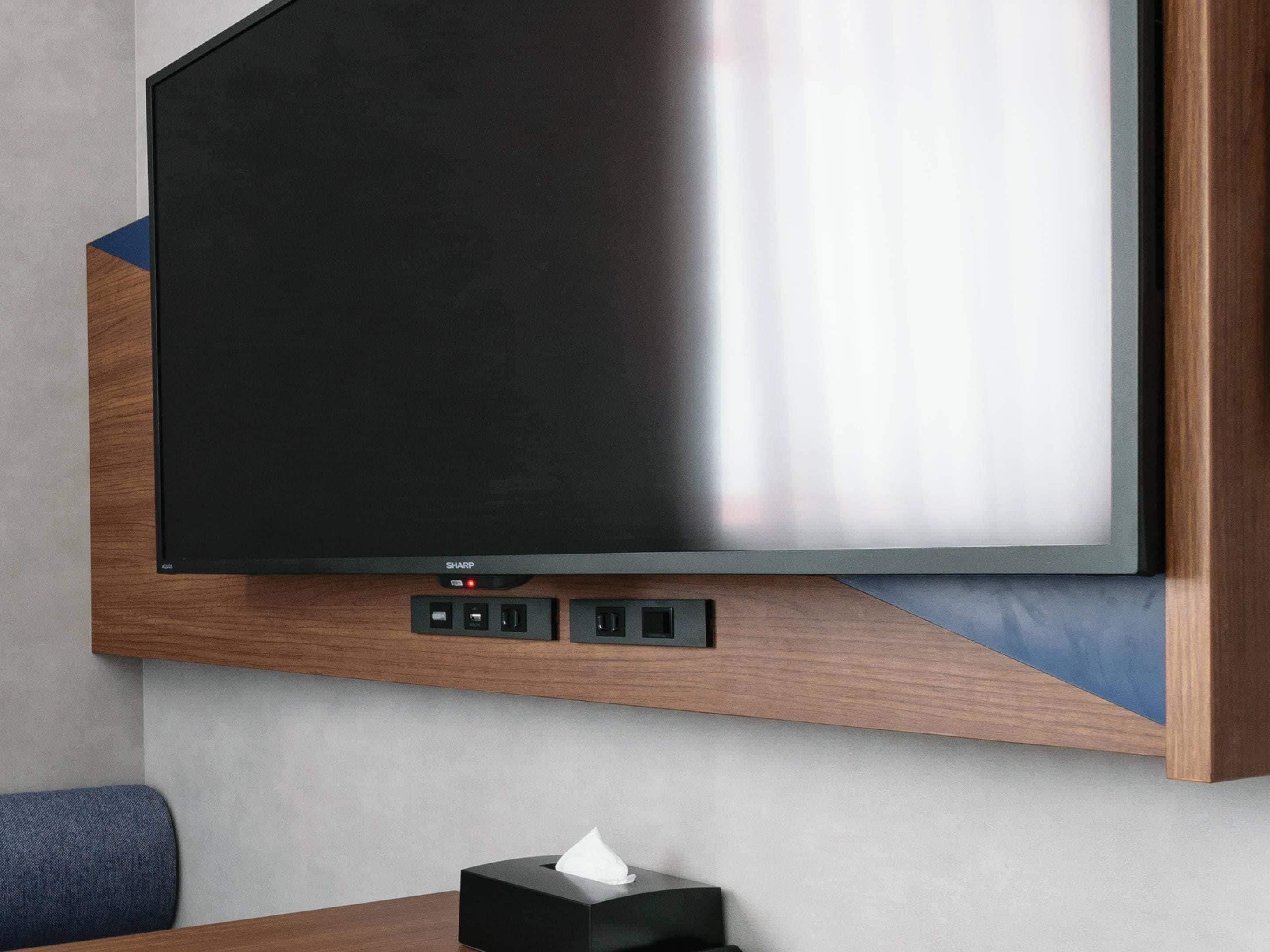 A modern hotel room with a large flat-screen TV mounted on a wooden wall, a small table with a tissue box, and a blue upholstered chair.