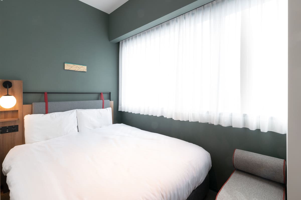 A neatly made bed in a room with a large window covered by sheer curtains.