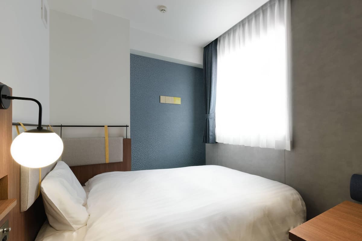 A hotel room with a bed, a bedside table, a lamp, and a window with curtains.