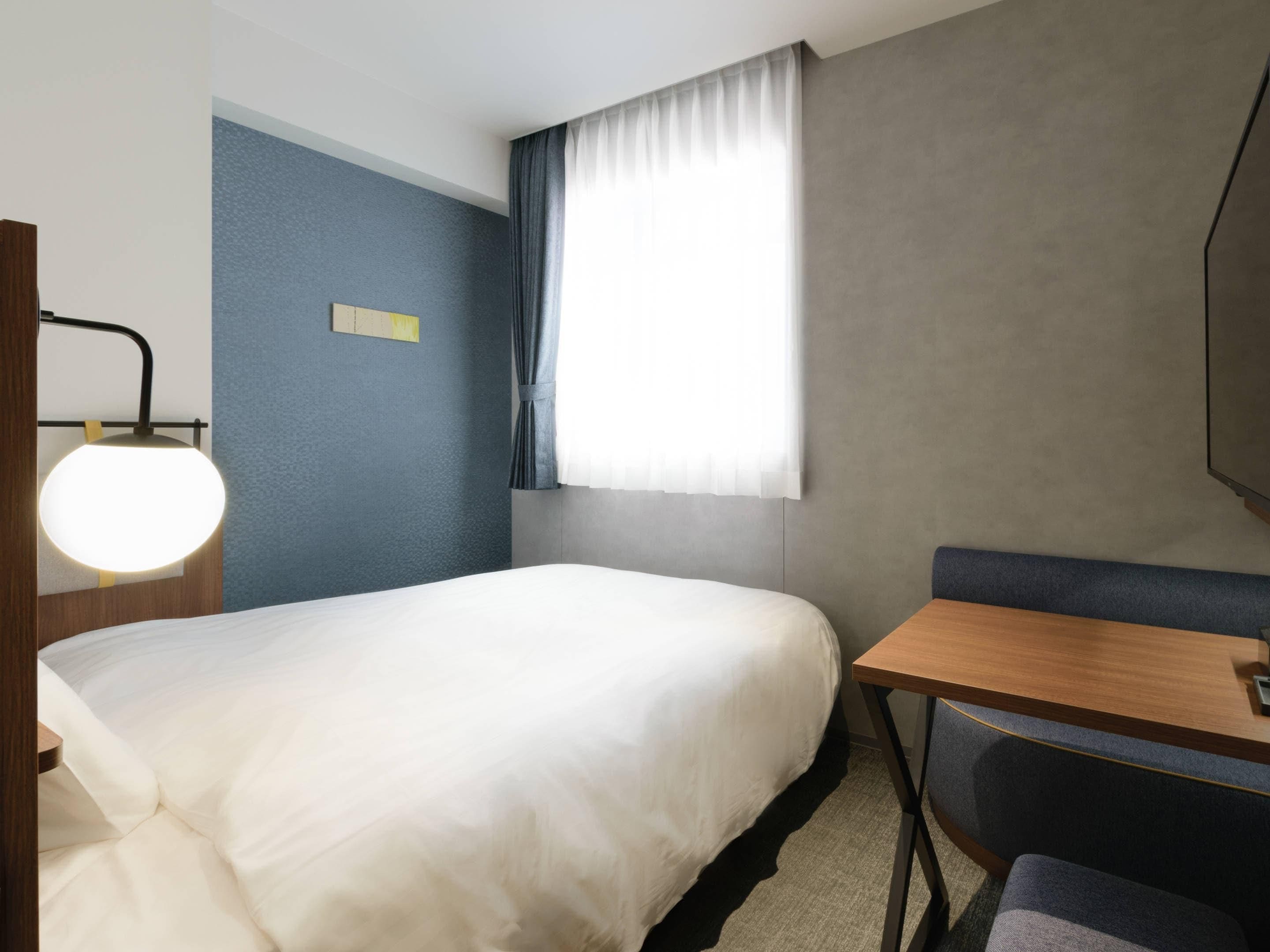 Ein Hotelzimmer mit Bett, Schreibtisch, Stuhl, Lampe und einem Fenster mit Vorhängen.