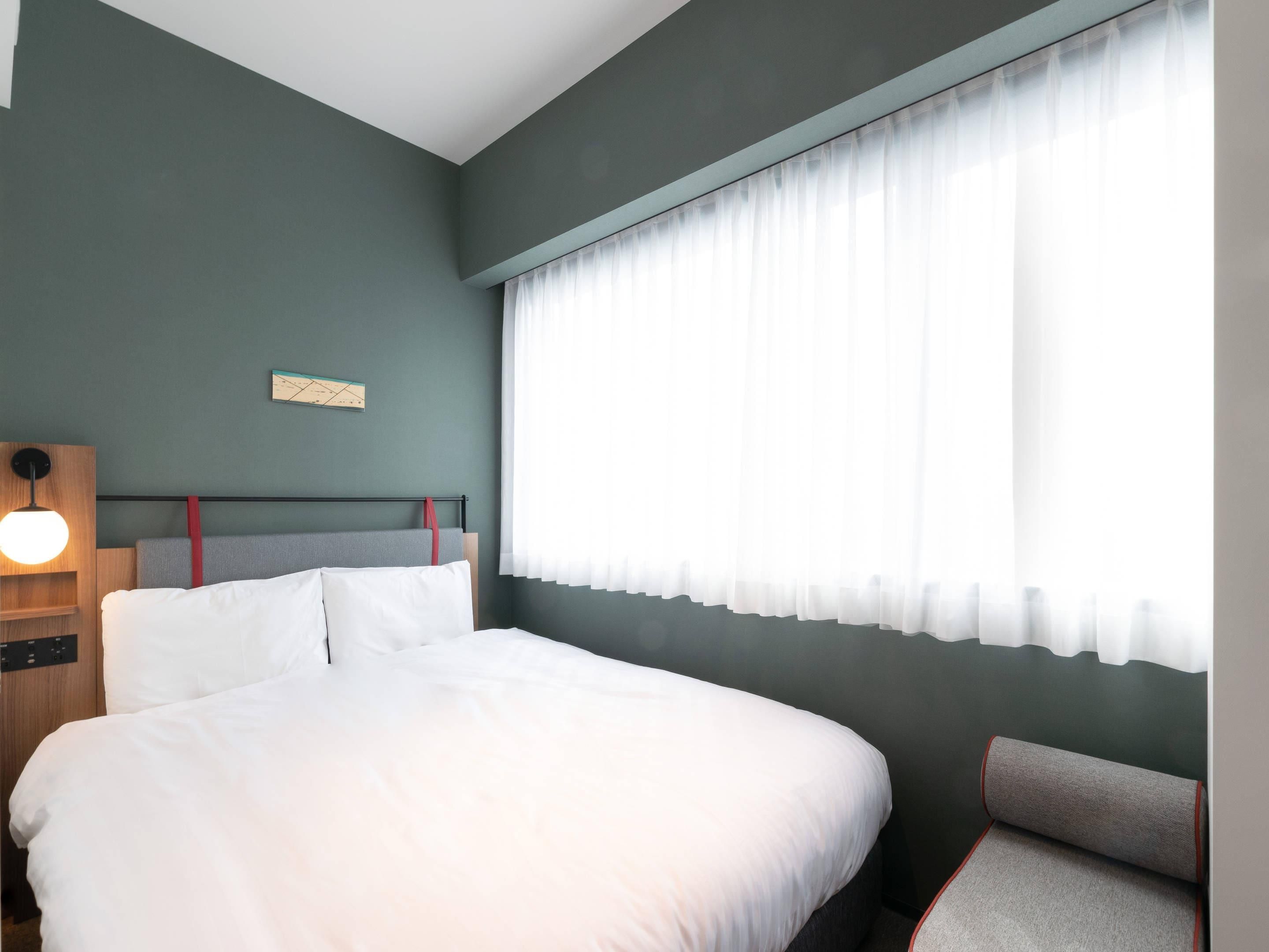 A modern hotel room with a large bed, a bedside table with a lamp, and a window with sheer curtains.