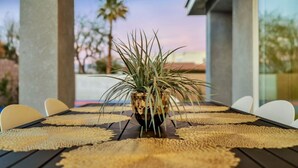A table set with woven placemats, a potted plant, and white chairs in an outdoor setting with a view of palm trees and a building. - Pool/Spa-Location-Basketball-GameRoom-Specials ask (Bermuda Dunes)