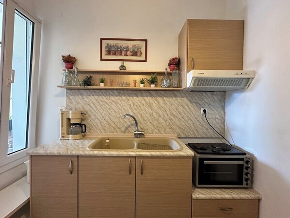 A compact kitchen with a sink, stove, and microwave. There is a coffee maker on the counter, and a small shelf with decorative items above the sink.