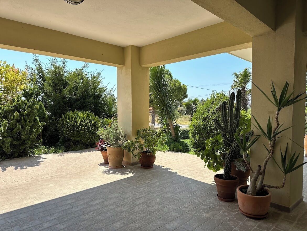 Un espacio al aire libre techado con piso de baldosas, plantas en macetas y vista a la vegetación circundante.