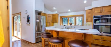 A modern kitchen with wooden cabinets, a stainless steel refrigerator, a large island with stools, and a built-in oven.