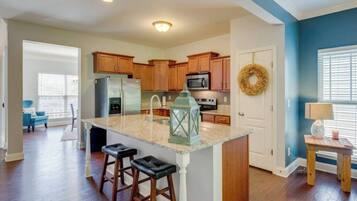 A modern kitchen with wooden cabinets, a stainless steel refrigerator, and a granite countertop. There are two bar stools and a lantern on the island.