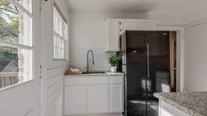Una cocina con muebles blancos, un refrigerador de acero inoxidable, una encimera de granito y una ventana con vista a una casa.