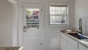 Un baño pequeño con paredes blancas, una ventana, un lavamanos con grifo y una puerta con un pequeño ventanal.