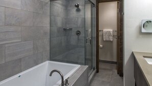 Une salle de bain moderne avec une douche à parois de verre, une baignoire blanche et une porte en bois donnant sur une autre salle de bain équipée d’un porte-serviettes.