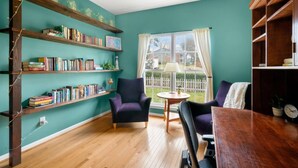 Un salón acogedor con paredes verde azulado, estantes de madera llenos de libros, dos sillones morados, una mesa de centro de madera y vista a una valla blanca de piquete.