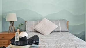 Un dormitorio con una cama, almohadas, una lámpara, un control remoto y una cesta con una manta.