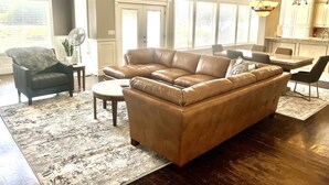 Ein Wohnzimmer mit braunem Ledersofa, einem dunklen Sessel, einem runden Couchtisch und einem Essbereich mit Tisch und Stühlen.