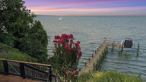 A wooden deck with a view of a calm body of water, a dock extending into the water, and a sailboat in the distance.