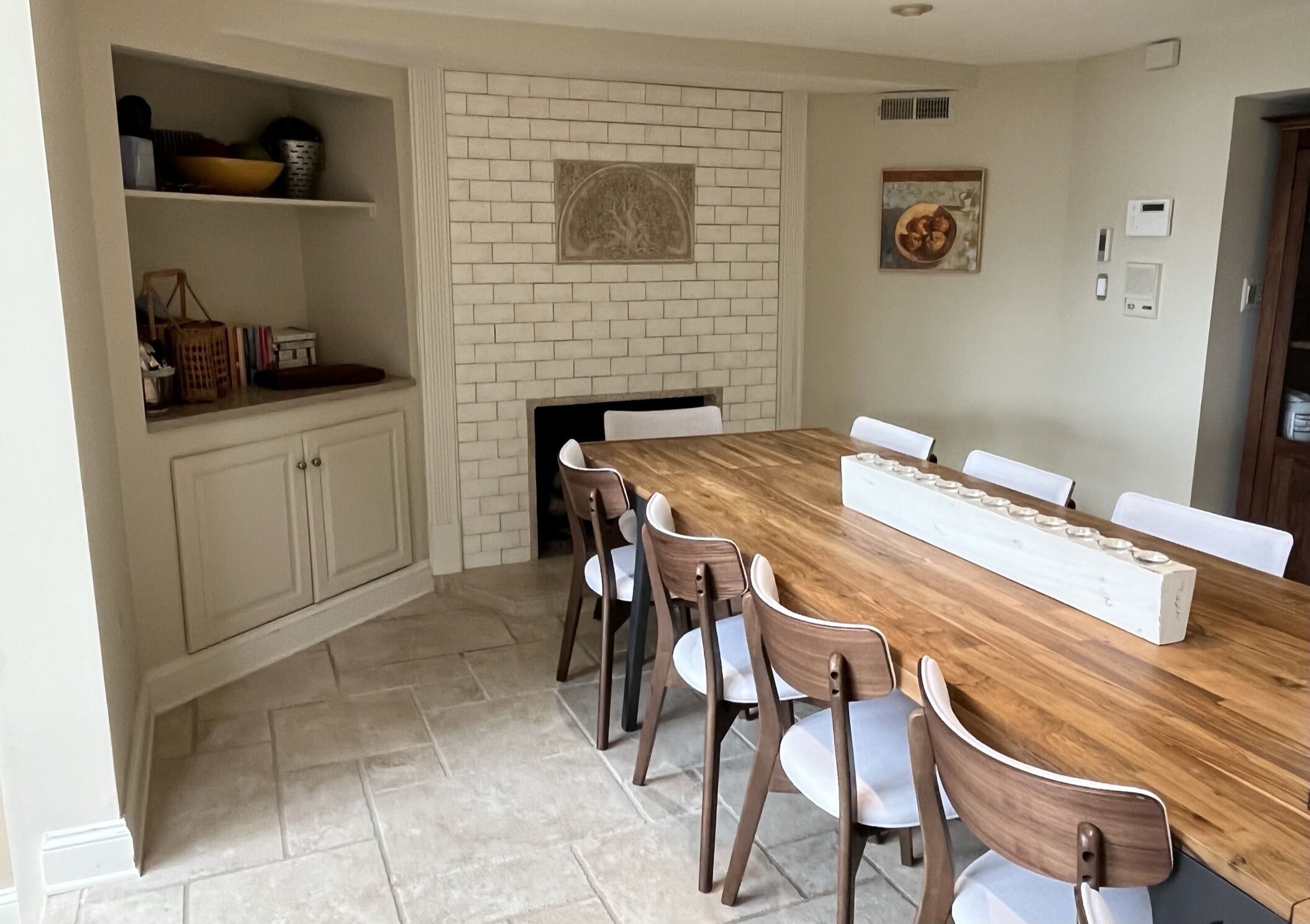 A dining room with a wooden table and chairs, a white brick fireplace, and a shelf with kitchen items.