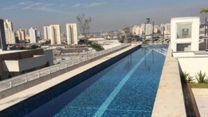 Ein langer, schmaler Swimmingpool mit glatter, blauer Wasseroberfläche, umgeben von einer niedrigen Betonmauer und einem Metallgeländer. Im Hintergrund sind moderne Gebäude und ein klarer Himmel zu sehen.