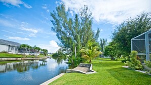 A serene waterfront property with a grassy lawn, a small bridge, and a clear sky. - Flamingo's Nest - A Tropical Waterfront Pool Oasis (Cape Coral)