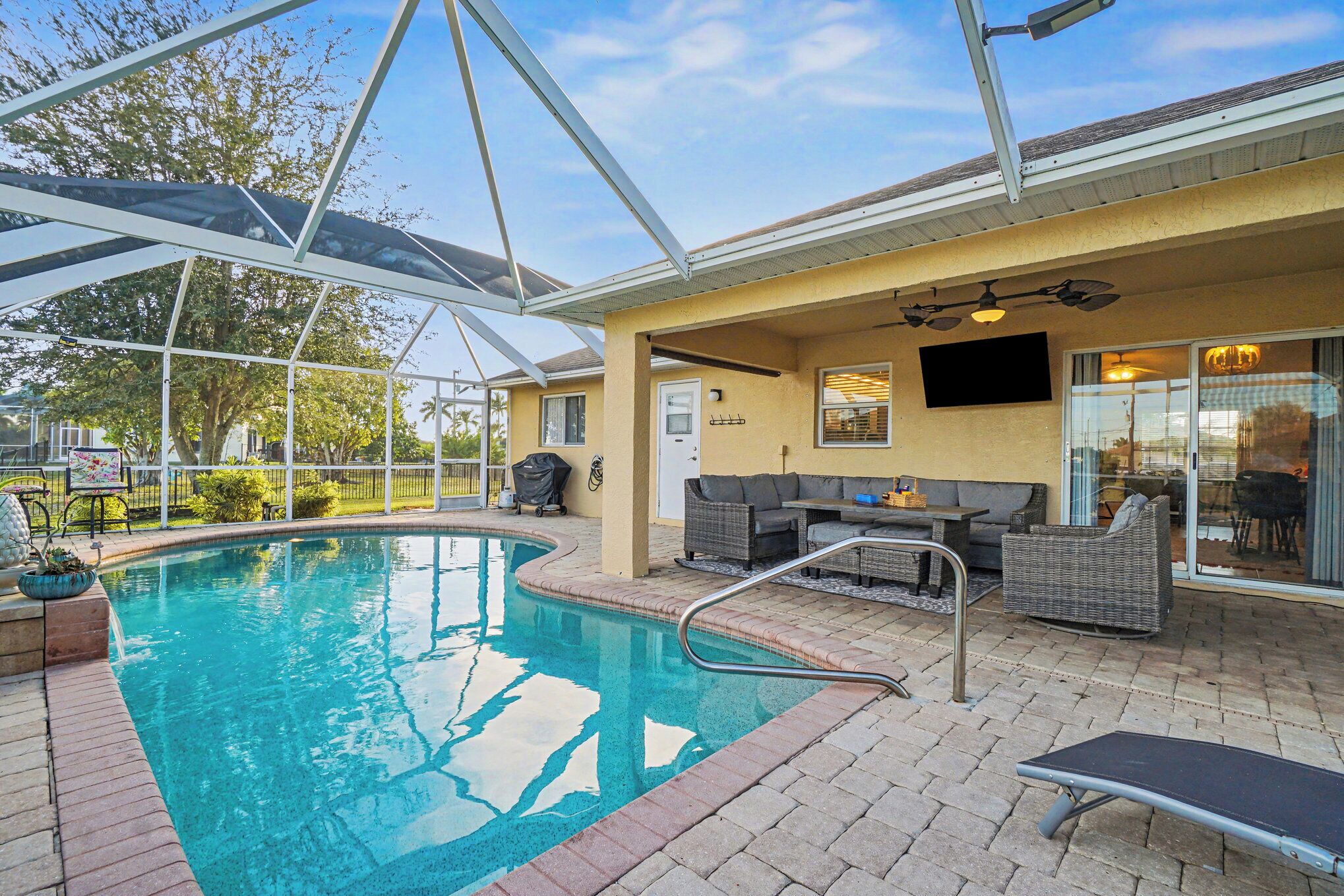 A covered outdoor swimming pool area with a patio, seating, and a television.