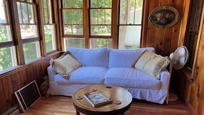Una habitación con piso de madera, un sofá blanco con cojines estampados, una mesa redonda con un libro y un posavasos, y un ventilador.