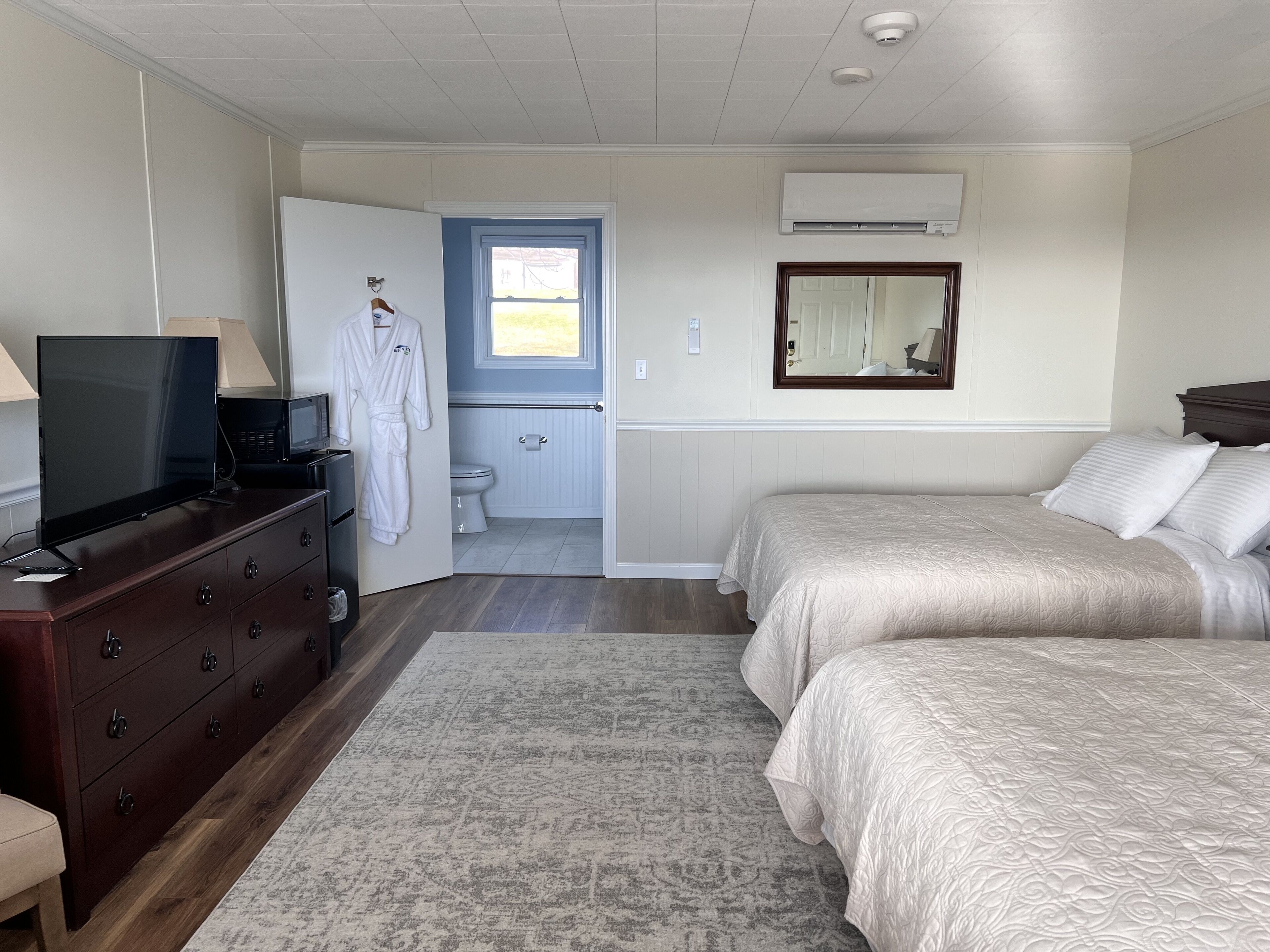 A bedroom with a TV on a dresser, a bathroom with a toilet and sink, and two beds with beige bedding.