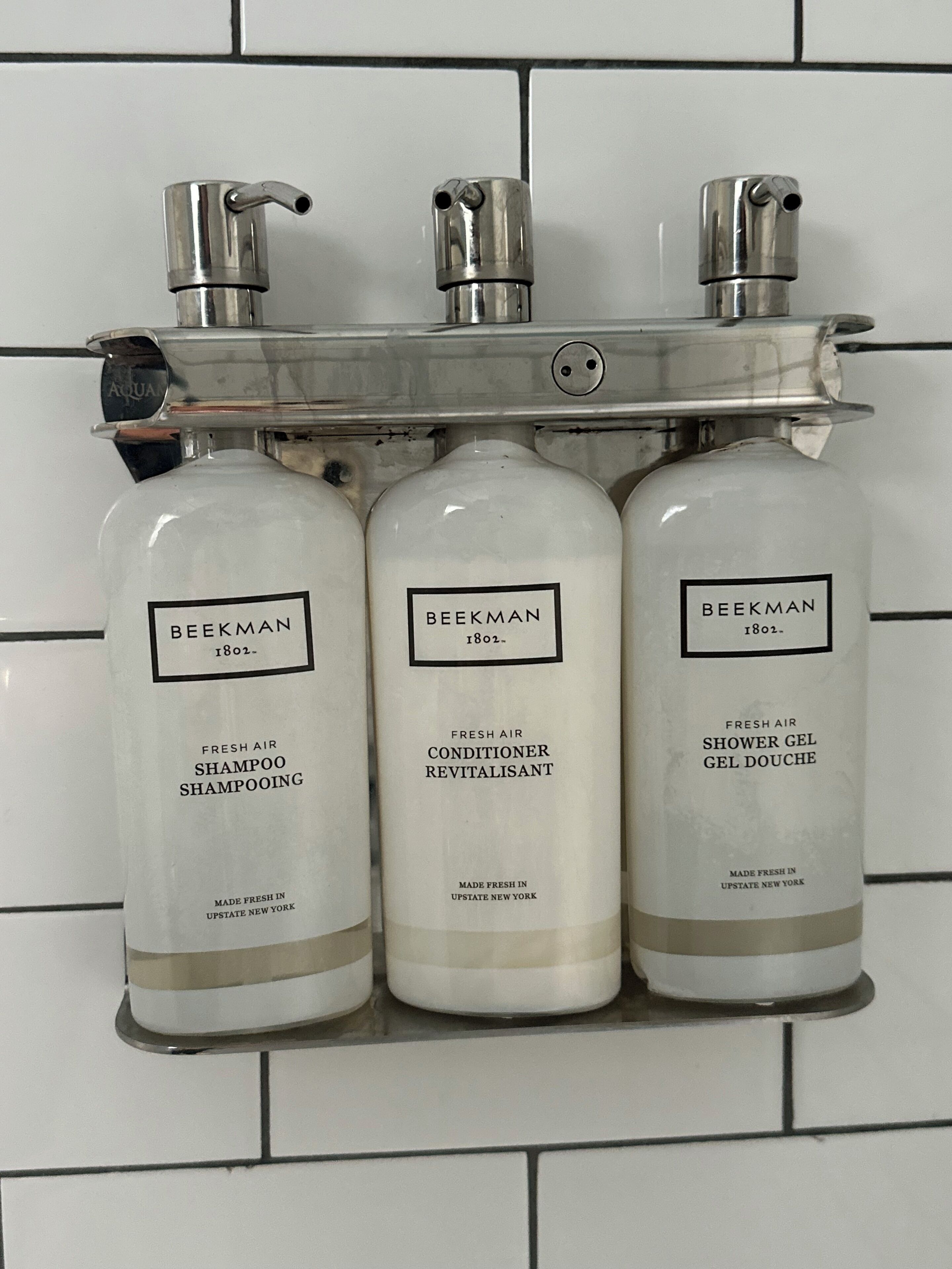 Three bottles of Beekman 1802 fresh air shampoo, conditioner, and shower gel on a white tiled wall.