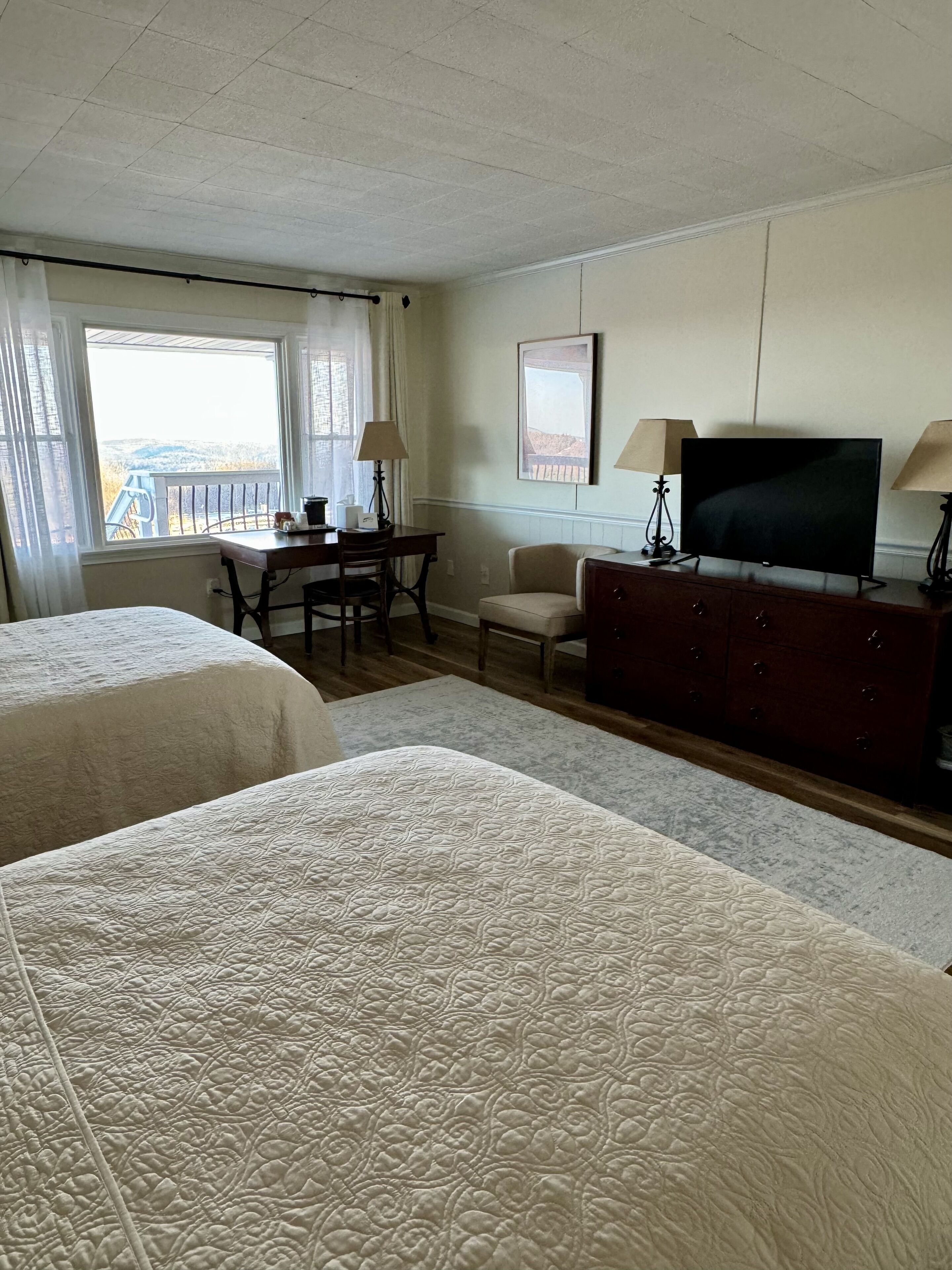 A hotel room with two beds, a desk with a chair, a television, and a view of the outdoors.