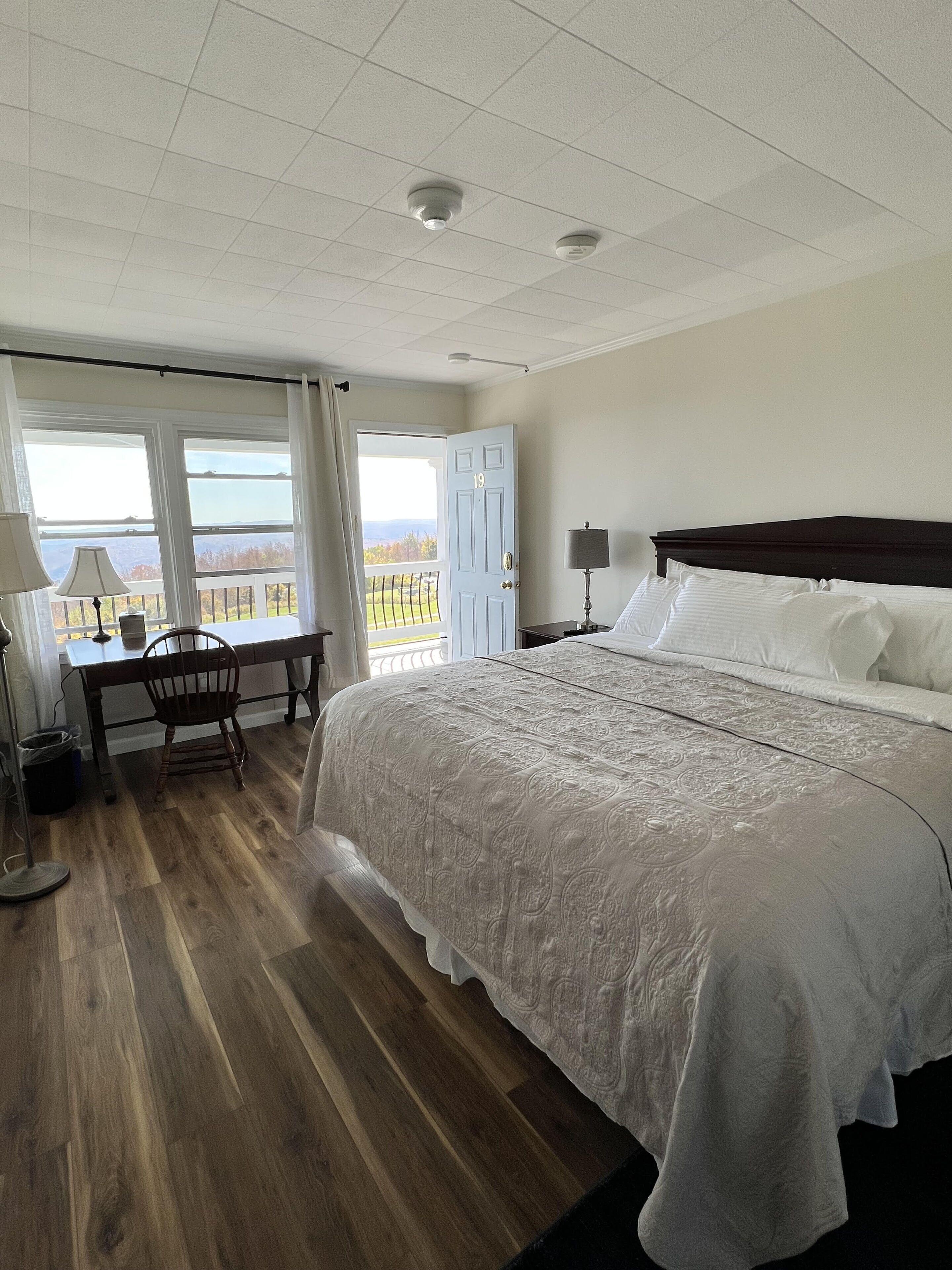A hotel room with a large bed, a desk, and a chair. There are windows with a view of the outdoors.