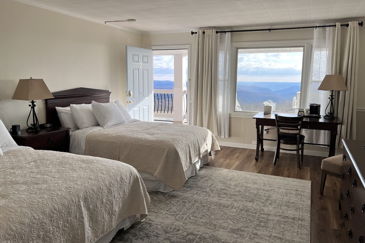A hotel room with two beds, a desk, a chair, a lamp, and a view of the mountains.
