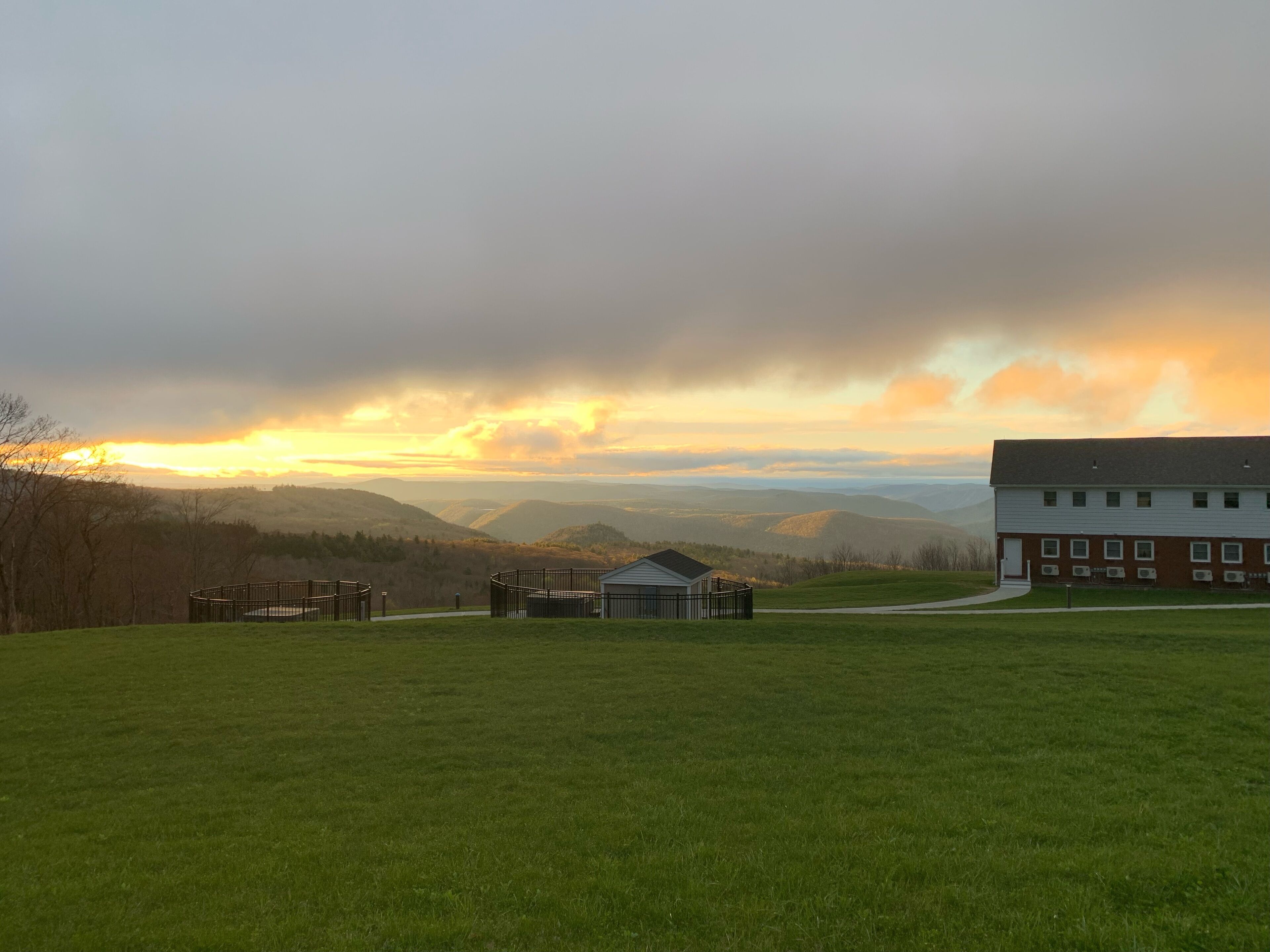 Ein grasbewachsenes Feld mit einem Gebäude und einem eingezäunten Bereich im Vordergrund, und eine gebirgige Landschaft im Hintergrund.