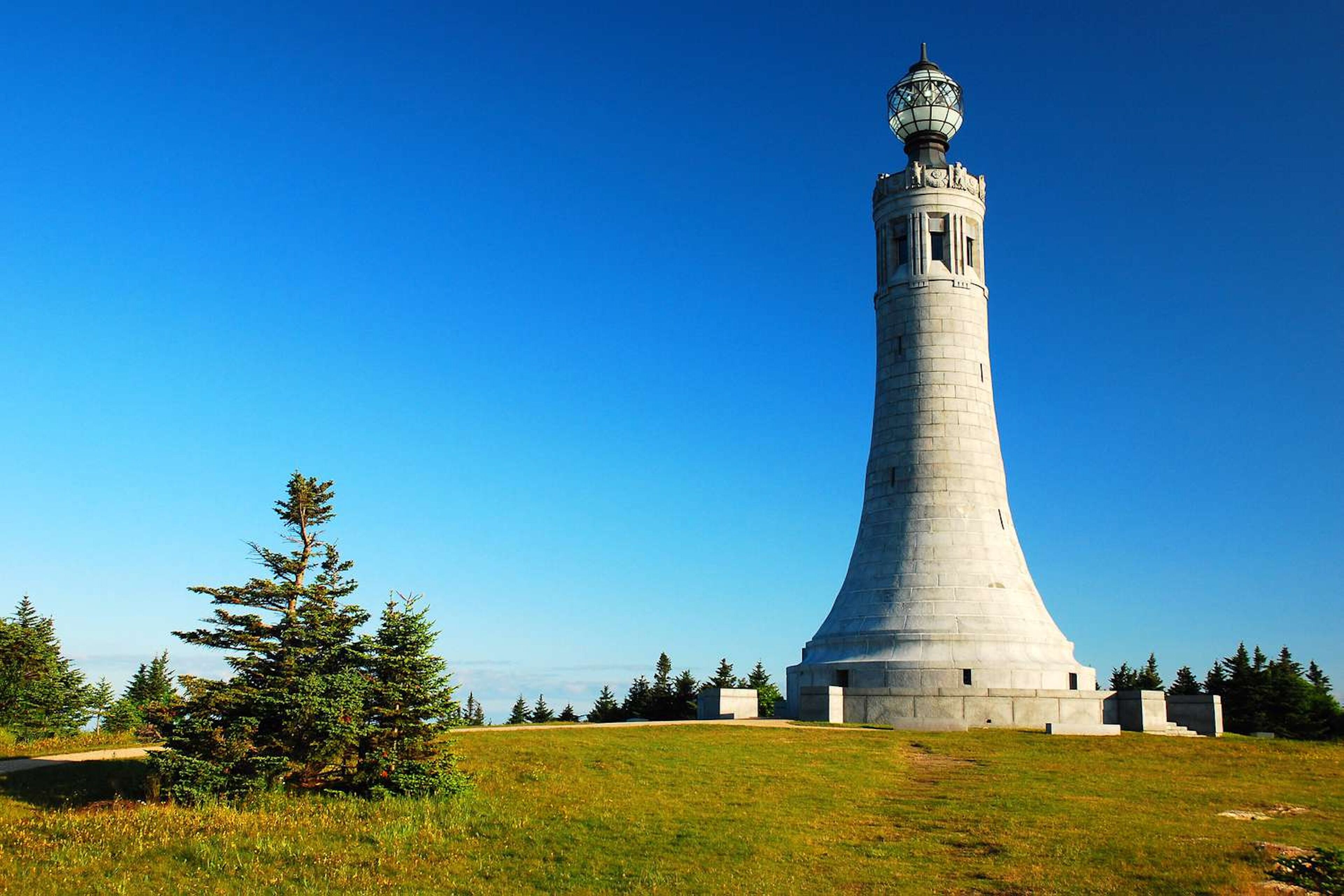 Un grand phare blanc, surmonté d’une sphère, entouré d’une zone herbeuse et d’arbres.