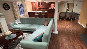A living room with a sectional sofa, a coffee table, and a patterned rug. There is a dining area with a table and chairs, and a kitchen in the background. - Bright and Clean Basement Apartment,5 min to Lakes. (Richmond Hill)