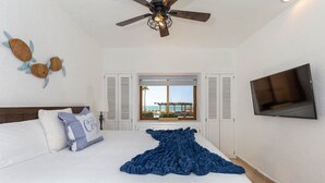 Un dormitorio con cama, ventilador de techo, televisor y ventana con vista a la playa.